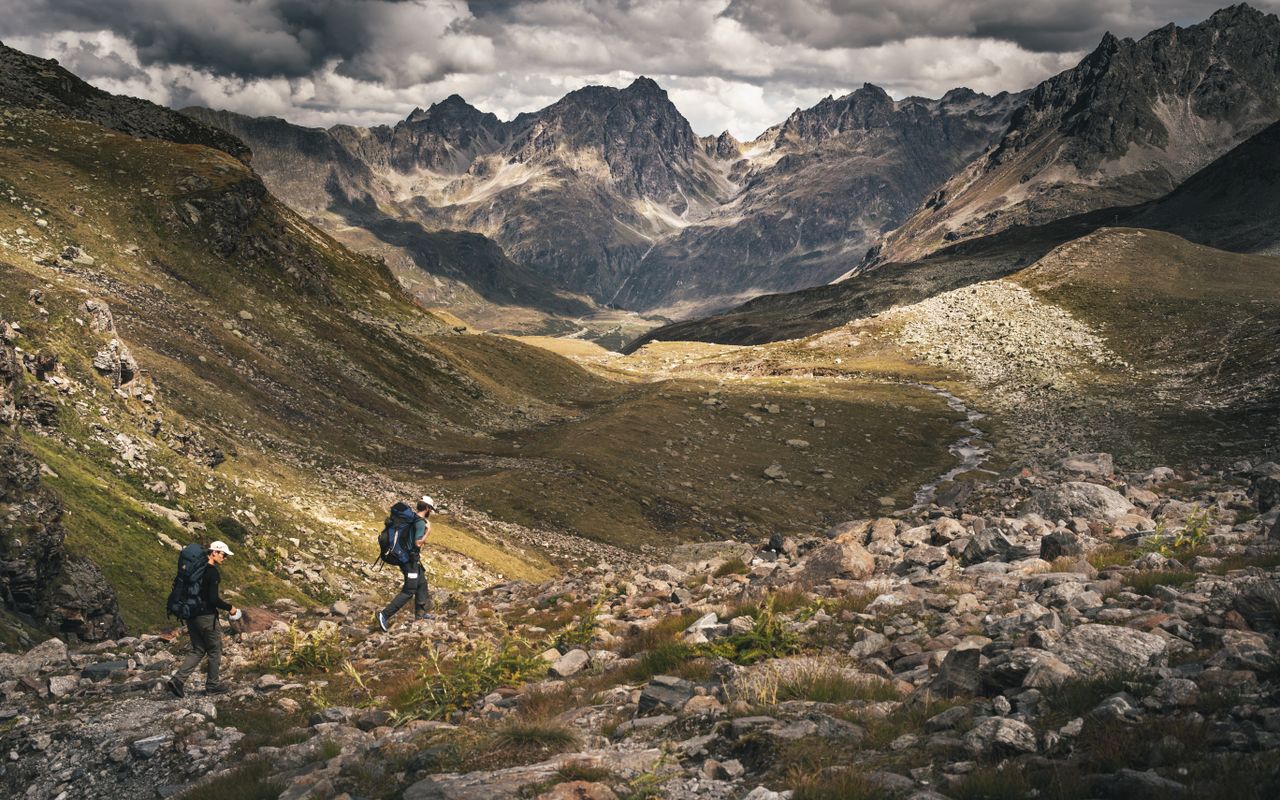 Two hikers explore the rocky trails of the Austrian Alps under a dramatic cloudy sky.