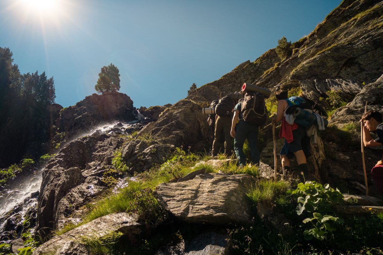 Hikers with backpacks climb a rocky mountain path under bright sun in CT, Spain.